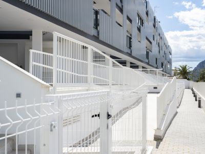 Exterior moderno de edificio de apartamentos con barandillas blancas, pasarelas pavimentadas y escaleras, con palmeras y montañas al fondo bajo cielo azul.