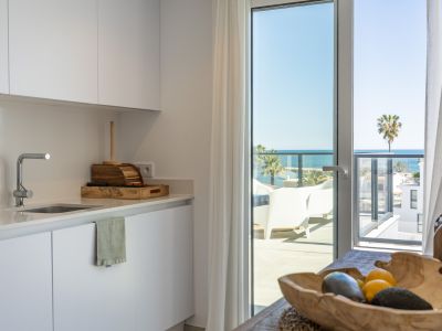Moderna cocina blanca con gabinetes elegantes y fregadero, iluminada por luz natural, con acceso directo a una terraza que ofrece vistas claras al mar y palmeras lejanas.