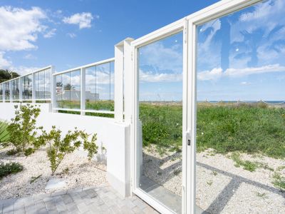 Moderna valla de cristal y puerta blanca que enmarca la vista a la vegetación costera natural y el mar Mediterráneo, con zona pavimentada y jardín de grava.