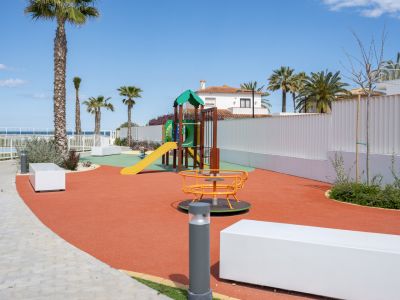 Parque infantil moderno con suelo de seguridad rojo, estructura de juegos colorida y carrusel, rodeado de palmeras y vistas lejanas al mar.