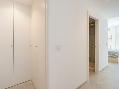 Pasillo con armarios empotrados blancos, suelo de gres claro, y vistas a un baño con ducha de azulejos oscuros y a un dormitorio con vistas al mar.