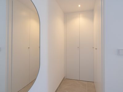 Pasillo minimalista con armarios empotrados blancos y tiradores discretos. Un espejo ovalado refleja la luz cálida del techo sobre el suelo de gres.