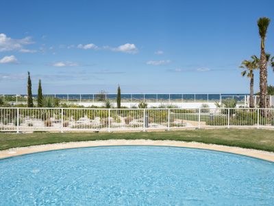 Piscina comunitaria con borde curvo, césped verde y jardín mediterráneo. Vistas al mar bajo un cielo azul claro.