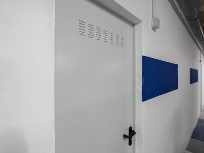 Puerta de trastero metálica blanca con rejillas de ventilación y maneta negra, en un pasillo de garaje con franjas azules.