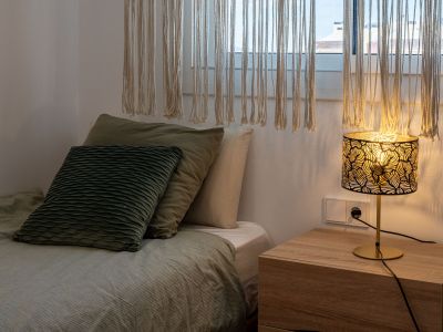 Rincón acogedor de dormitorio con cama vestida en tonos verdes, mesita de noche de madera clara y lámpara de diseño con pantalla calada. Cortinas de macramé filtran la luz natural.