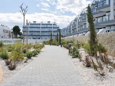 Sendero pavimentado rodeado de grava blanca, plantas mediterráneas y luces de bolardo modernas, que conduce a edificios de apartamentos con balcones de cristal y muro de piedra natural.