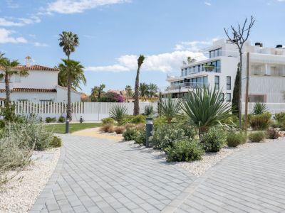Senderos adoquinados con bordes de grava blanca serpentean entre exuberante vegetación mediterránea, con palmeras y agaves, hacia edificios de apartamentos modernos y una casa tradicional bajo un cielo azul claro.