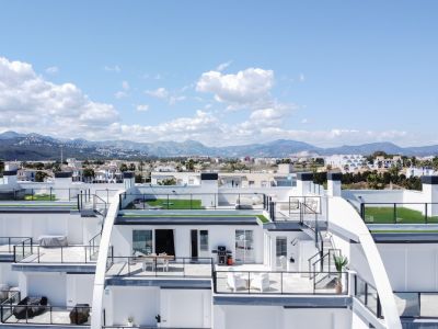 Vista aérea de apartamentos modernos de varios niveles con amplias terrazas, barandales de cristal, césped artificial y mobiliario exterior, con montañas y ciudad al fondo.