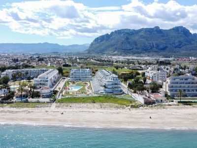 Vista aérea de complejos residenciales modernos con piscinas y palmeras, ubicados frente a una playa de arena y con la imponente montaña de Dénia al fondo.