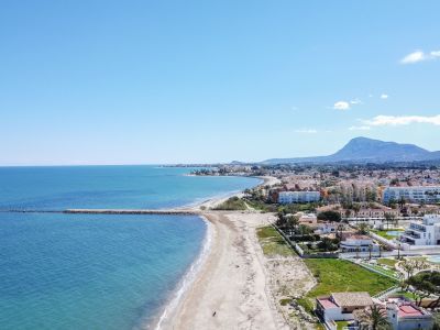 Vista aérea de la playa de arena de Dénia y el mar Mediterráneo turquesa, con un espigón de roca y edificios costeros, y el Montgó al fondo.