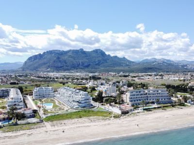Vista aérea de modernos apartamentos blancos junto a una playa de arena dorada y el mar turquesa, con la imponente sierra del Montgó al fondo.