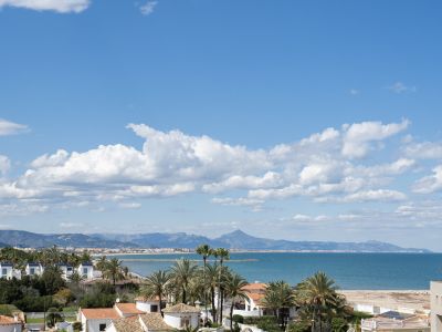 Vista panorámica de la costa mediterránea con edificios residenciales, palmeras, una playa de arena y montañas lejanas bajo un cielo azul con nubes blancas.