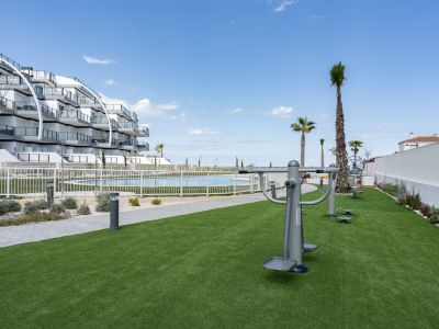 Zona de fitness exterior con césped artificial y equipamiento, junto a la piscina comunitaria y el moderno edificio de apartamentos, con vistas al mar.