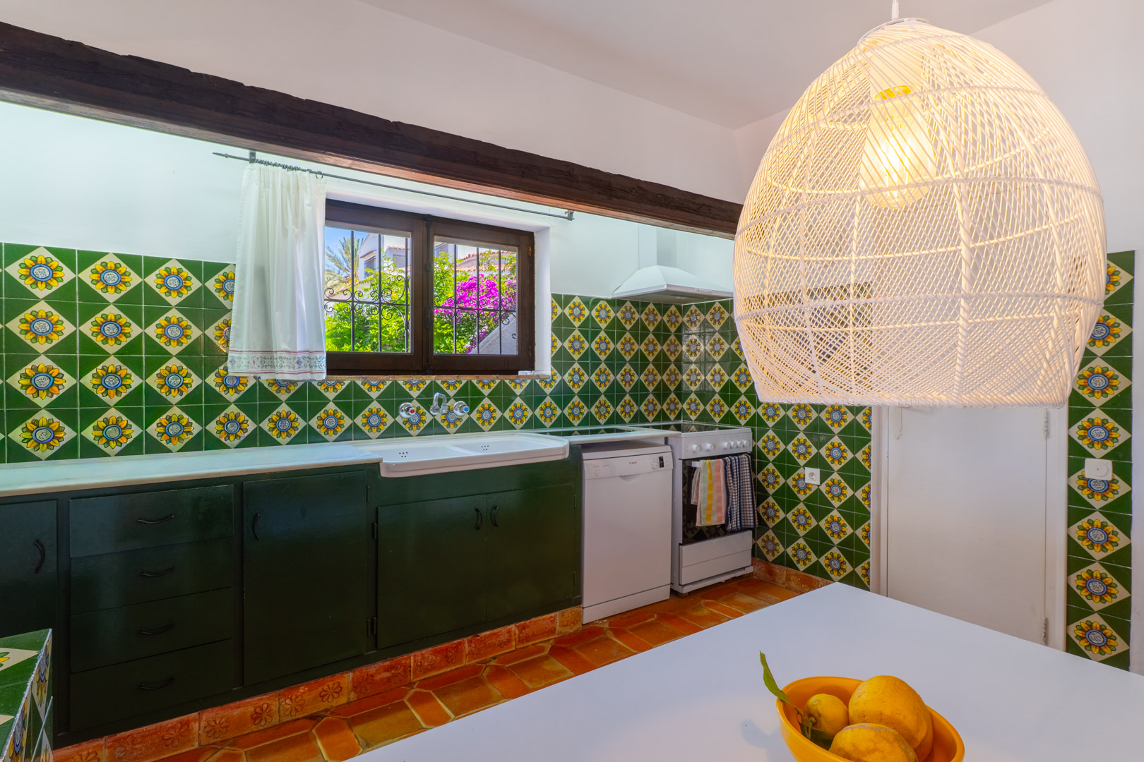 Cocina con azulejos decorativos, muebles verde oscuro y lámpara colgante de mimbre.