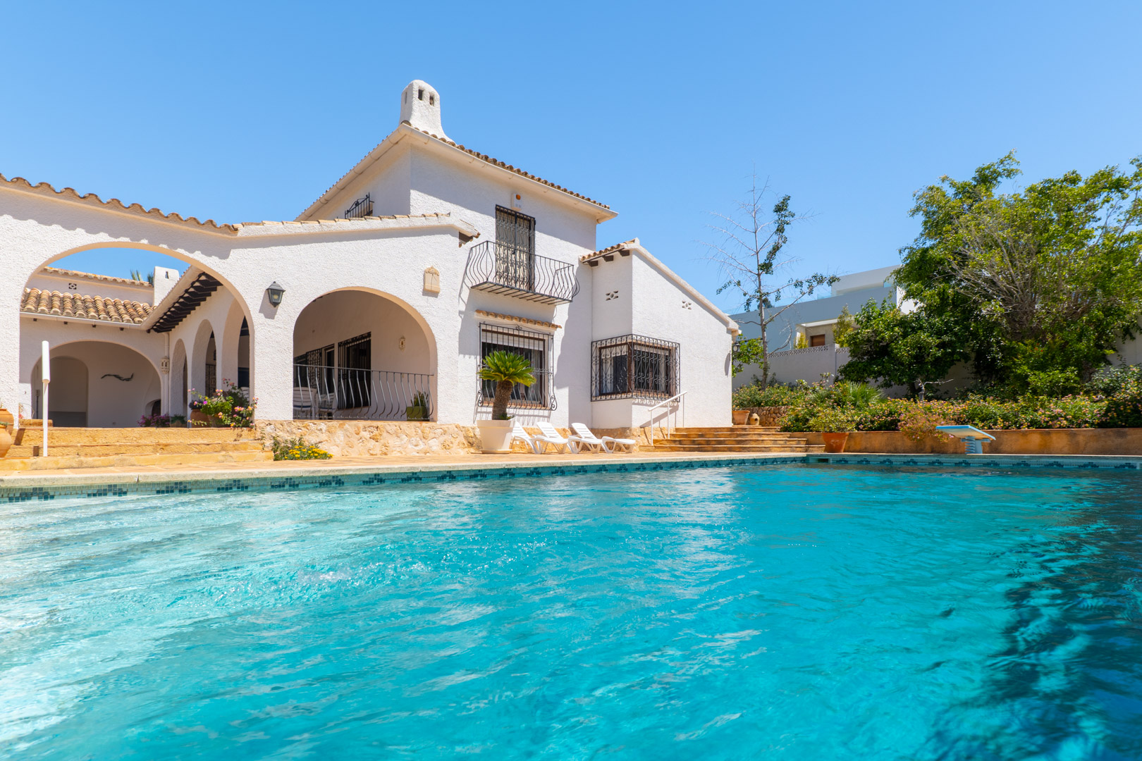 Piscina turquesa con borde de mosaico frente a la fachada blanca.