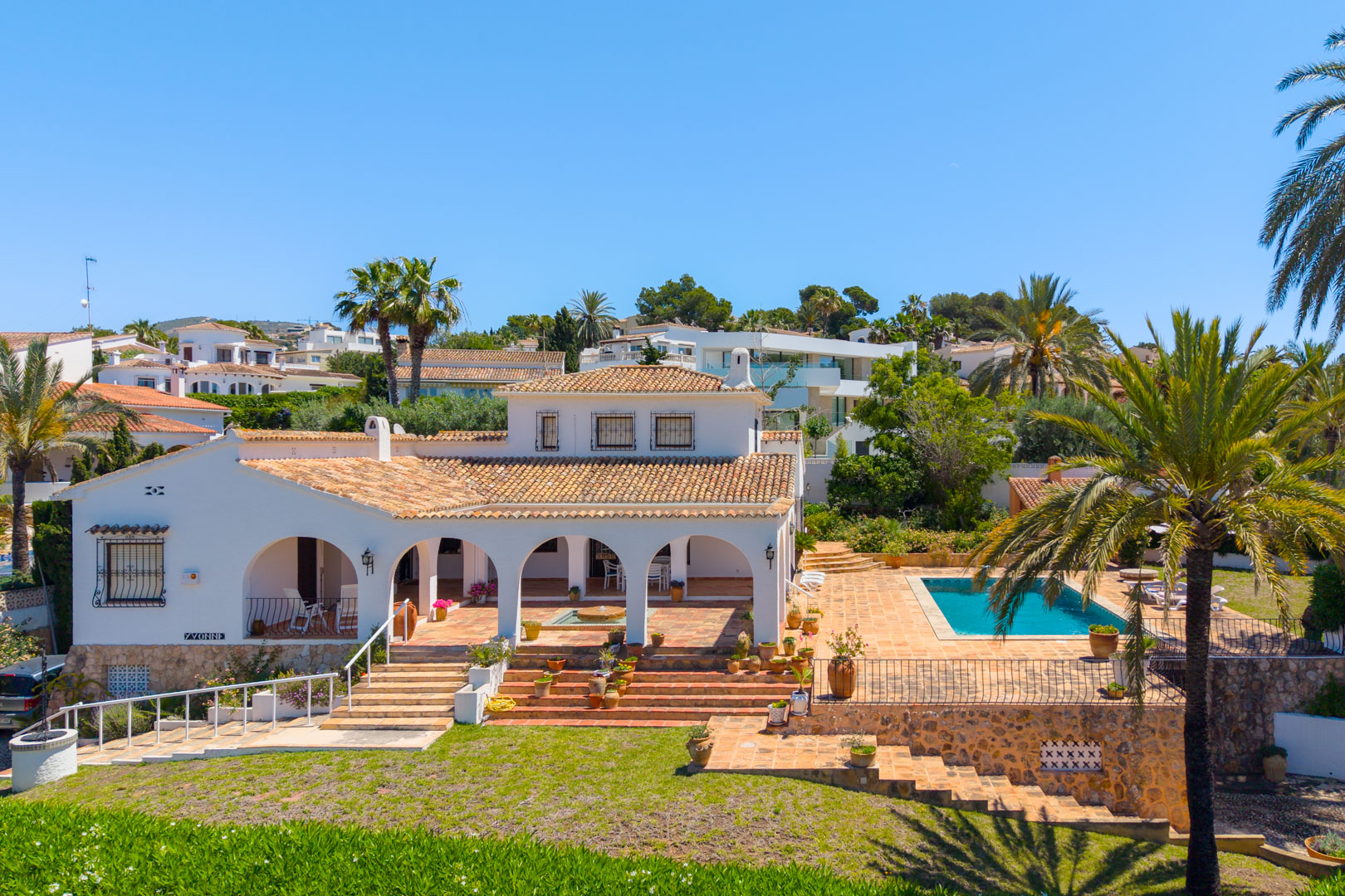 Vista elevada de la villa con arcos, piscina y jardín escalonado con vegetación.