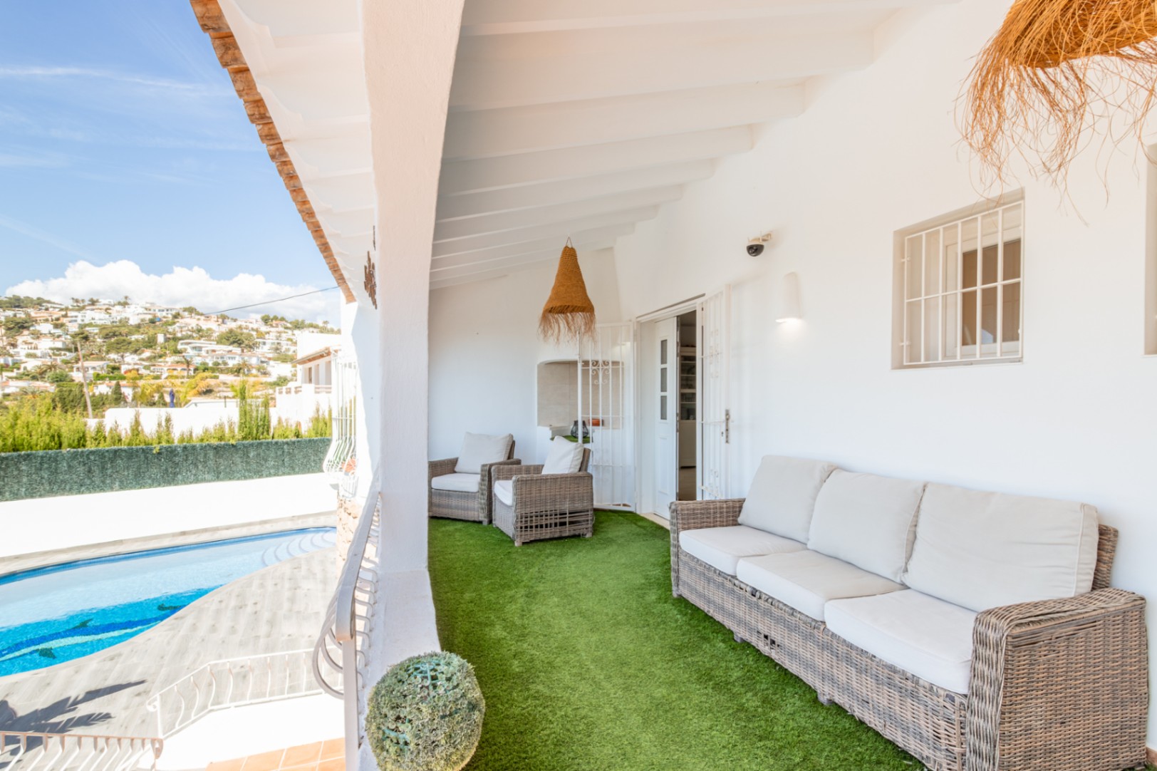 Amplia terraza cubierta con cómodos muebles de mimbre sobre césped artificial, ofreciendo vistas a la piscina privada y a las colinas residenciales bajo un cielo despejado. Amplia terraza cubierta con cómodos muebles de mimbre sobre césped artificial, ofreciendo vistas a la piscina privada y a las colinas residenciales bajo un cielo despejado.