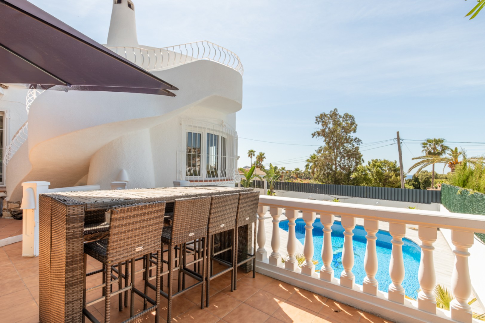Amplia terraza exterior con barra de ratán y taburetes, con vistas a la piscina privada. Se aprecia la singular arquitectura espiral de la villa blanca bajo un cielo despejado. Amplia terraza exterior con barra de ratán y taburetes, con vistas a la piscina privada. Se aprecia la singular arquitectura espiral de la villa blanca bajo un cielo despejado.