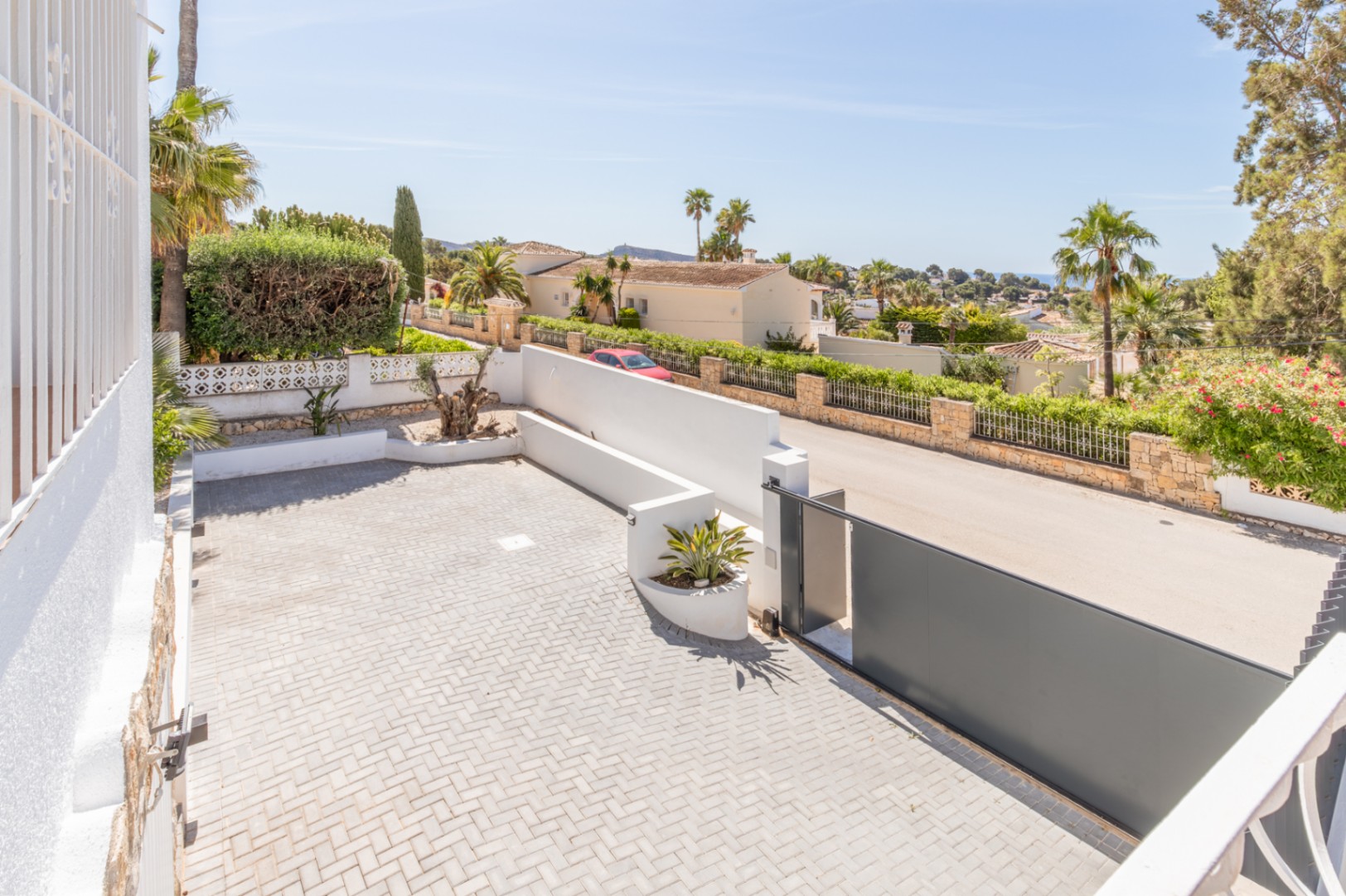 Amplia zona de aparcamiento pavimentada con adoquines en espiga y una moderna puerta corredera gris oscuro. Vistas a la zona residencial, vegetación mediterránea y el mar a lo lejos. Amplia zona de aparcamiento pavimentada con adoquines en espiga y una moderna puerta corredera gris oscuro. Vistas a la zona residencial, vegetación mediterránea y el mar a lo lejos.
