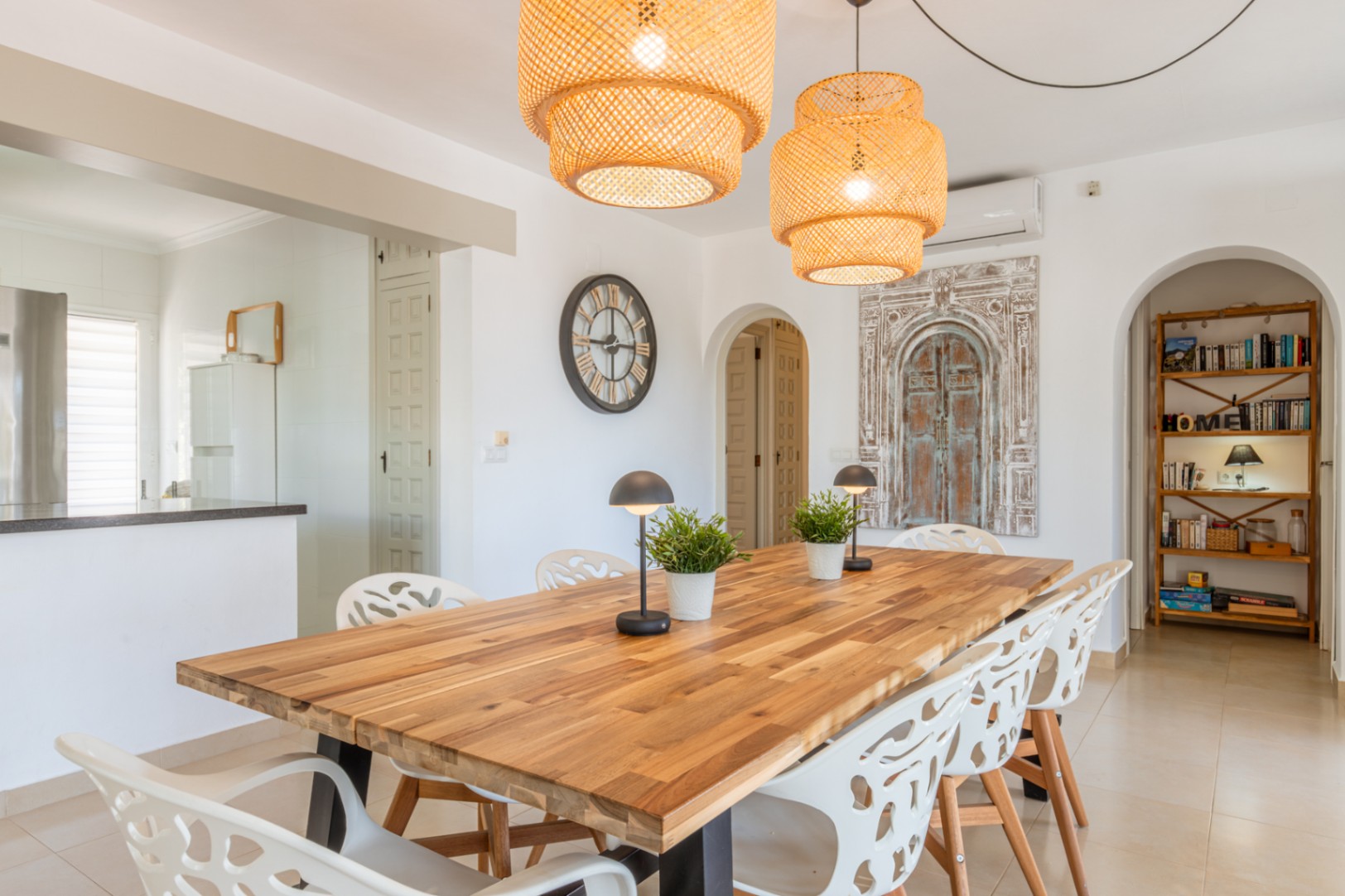Amplio comedor con mesa de madera maciza, sillas blancas modernas y dos lámparas colgantes de mimbre. Arcos conectan con la cocina y un rincón de lectura con estantería. Amplio comedor con mesa de madera maciza, sillas blancas modernas y dos lámparas colgantes de mimbre. Arcos conectan con la cocina y un rincón de lectura con estantería.