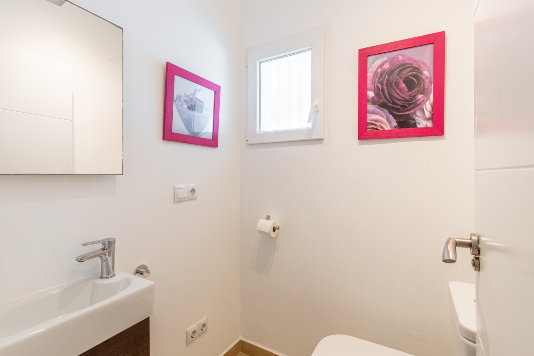 Aseo de cortesía con lavabo rectangular y grifería cromada. Destacan los detalles decorativos con marcos fucsia y una ventana que aporta luz natural. Aseo de cortesía con lavabo rectangular y grifería cromada. Destacan los detalles decorativos con marcos fucsia y una ventana que aporta luz natural.