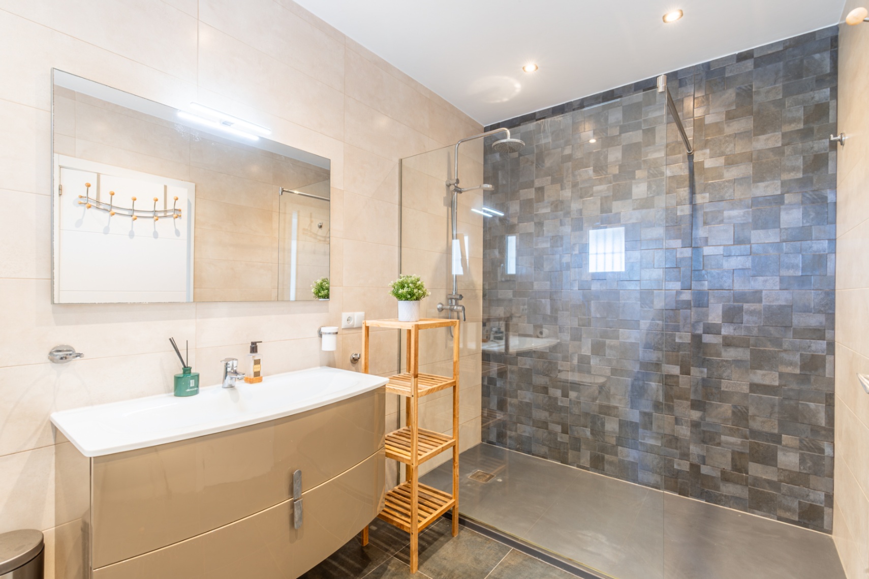 Baño moderno con lavabo de mueble beige brillante, espejo iluminado y ducha de obra con mampara de cristal y azulejos mosaico grises. Suelos oscuros y paredes claras. Baño moderno con lavabo de mueble beige brillante, espejo iluminado y ducha de obra con mampara de cristal y azulejos mosaico grises. Suelos oscuros y paredes claras.