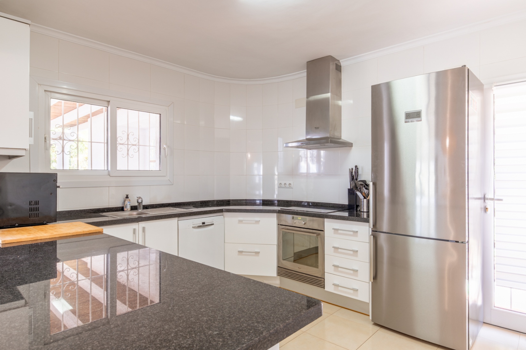 Cocina equipada con armarios blancos, encimeras de granito oscuro y electrodomésticos de acero inoxidable, incluyendo nevera y horno integrados. Ventana con rejas de seguridad que aporta luz natural. Cocina equipada con armarios blancos, encimeras de granito oscuro y electrodomésticos de acero inoxidable, incluyendo nevera y horno integrados. Ventana con rejas de seguridad que aporta luz natural.