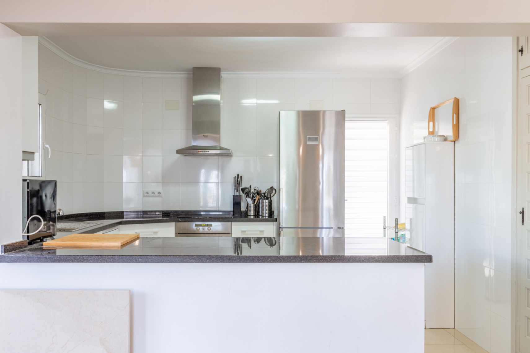 Cocina moderna y luminosa con paredes de azulejos blancos, encimeras de granito oscuro y electrodomésticos de acero inoxidable, incluyendo nevera y campana extractora. Dispone de una barra funcional. Cocina moderna y luminosa con paredes de azulejos blancos, encimeras de granito oscuro y electrodomésticos de acero inoxidable, incluyendo nevera y campana extractora. Dispone de una barra funcional.