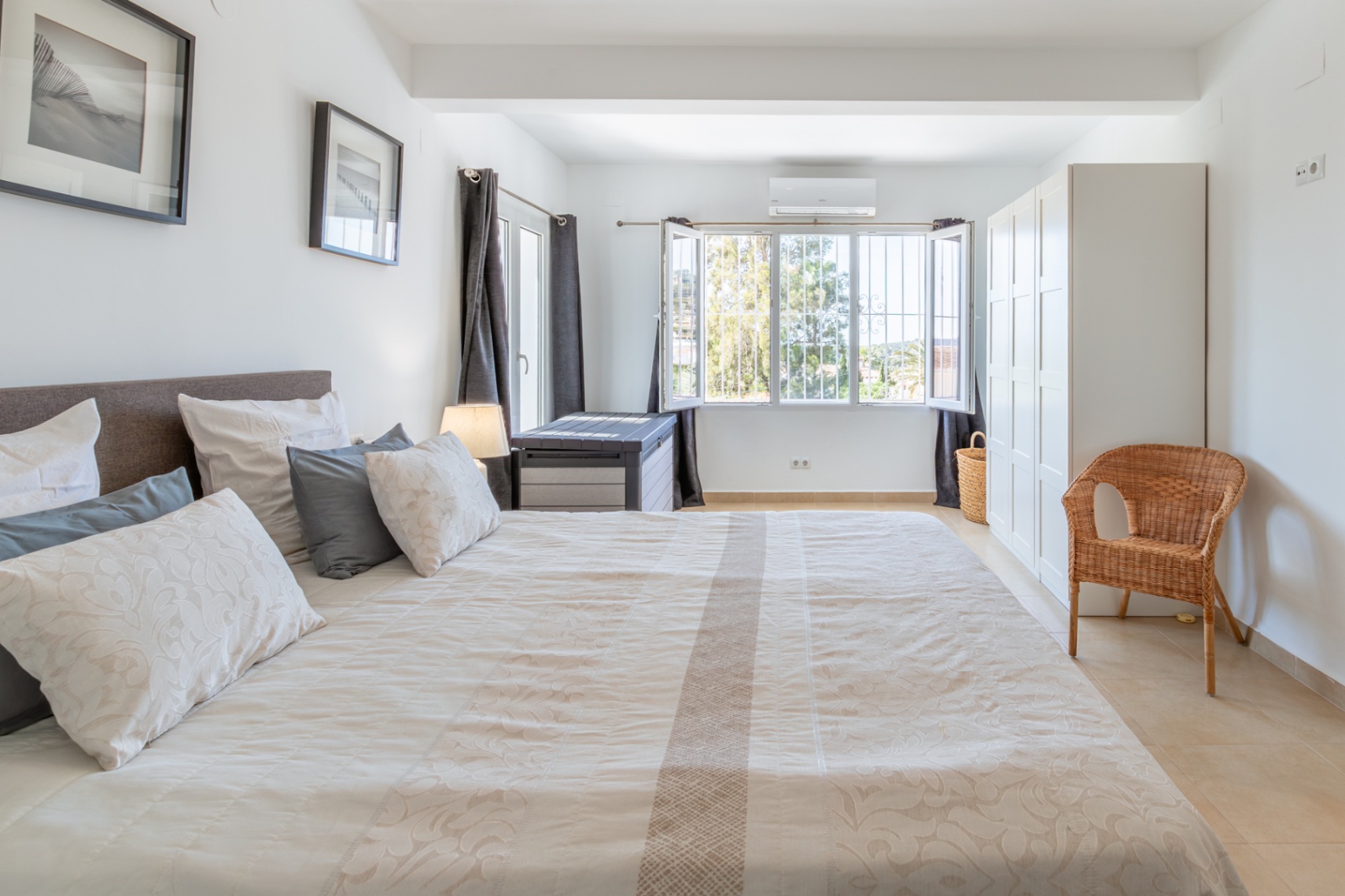Dormitorio principal con cama doble, suelo de baldosas claras, armario blanco y ventana con rejas, ofreciendo vistas al exterior y aire acondicionado. Dormitorio principal con cama doble, suelo de baldosas claras, armario blanco y ventana con rejas, ofreciendo vistas al exterior y aire acondicionado.