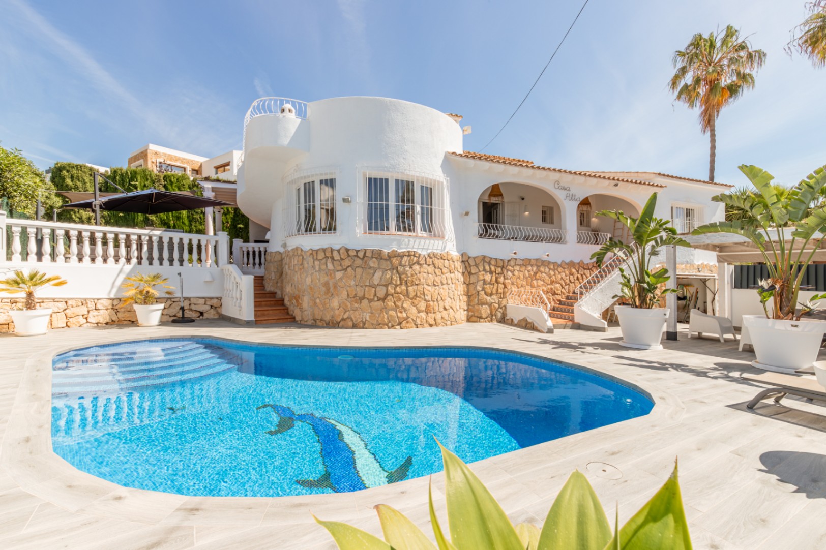 Exterior de villa blanca con piscina privada de mosaico azul y delfín. Destaca la base de piedra y una sección arquitectónica redonda con ventanas. Exterior de villa blanca con piscina privada de mosaico azul y delfín. Destaca la base de piedra y una sección arquitectónica redonda con ventanas.