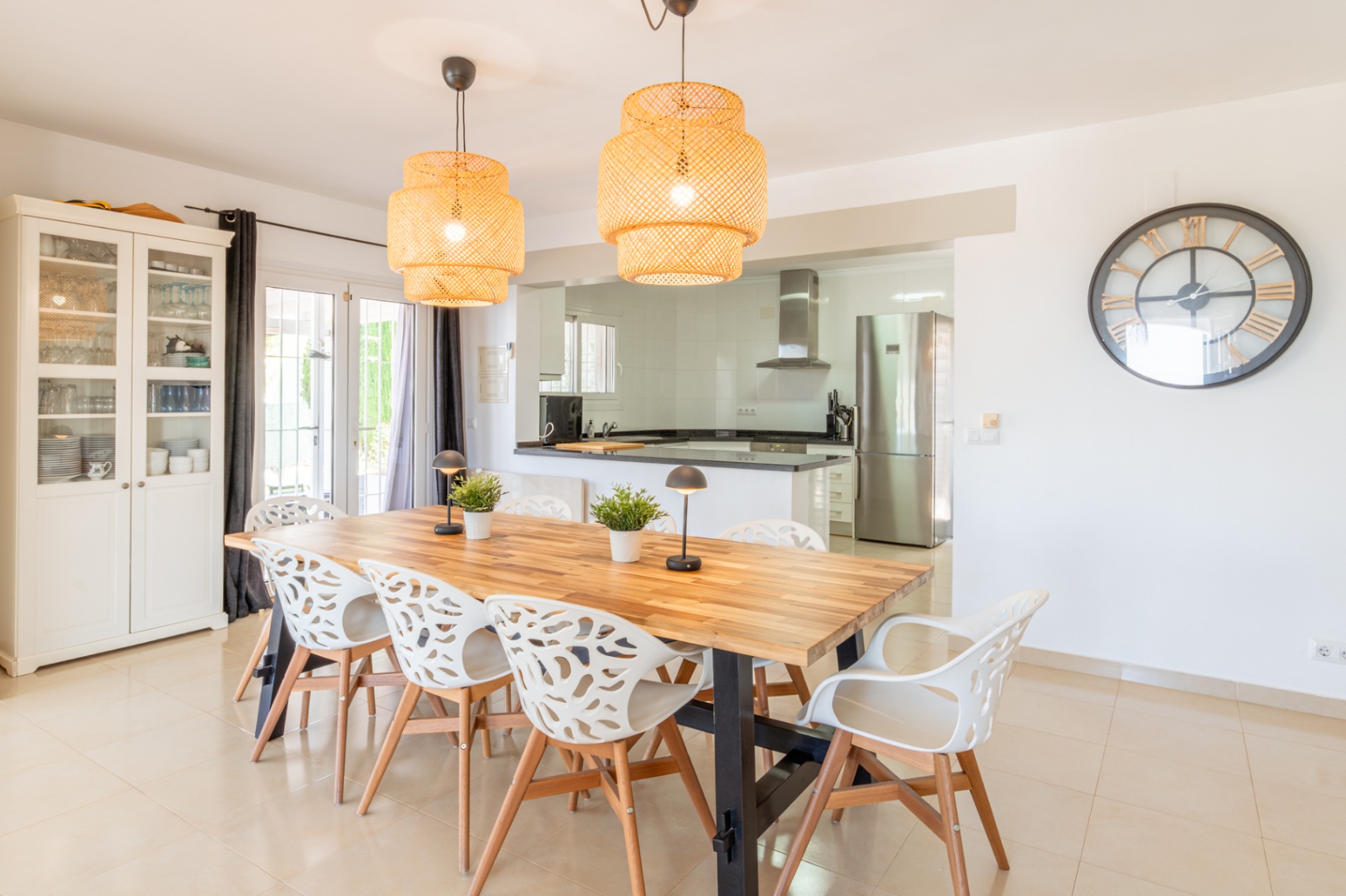 Luminoso comedor con una gran mesa de madera, ocho sillas blancas de diseño y dos lámparas colgantes de mimbre. Se conecta fluidamente a una cocina moderna y equipada con electrodomésticos de acero. Luminoso comedor con una gran mesa de madera, ocho sillas blancas de diseño y dos lámparas colgantes de mimbre. Se conecta fluidamente a una cocina moderna y equipada con electrodomésticos de acero.