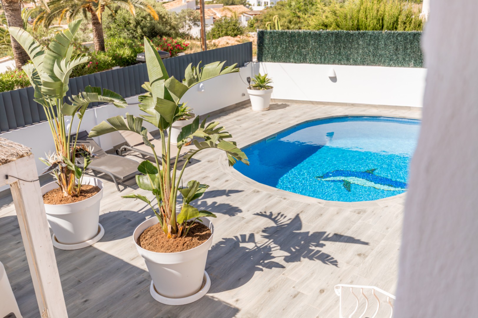Piscina privada de forma irregular con mosaico de delfín, rodeada por una terraza de baldosas imitación madera y plantas tropicales en macetas. Piscina privada de forma irregular con mosaico de delfín, rodeada por una terraza de baldosas imitación madera y plantas tropicales en macetas.