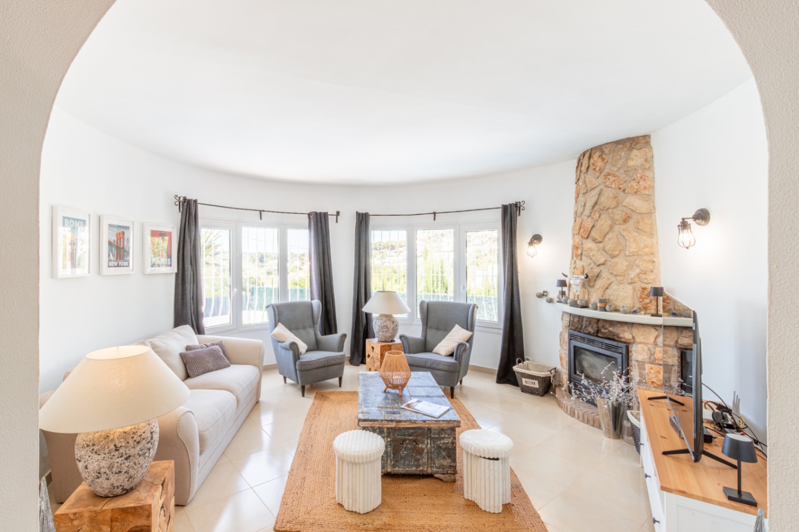 Salón con pared curva de ventanales que aportan luz natural, chimenea revestida de piedra, y suelos de baldosas claras. Alfombra de yute y mobiliario confortable. Salón con pared curva de ventanales que aportan luz natural, chimenea revestida de piedra, y suelos de baldosas claras. Alfombra de yute y mobiliario confortable.