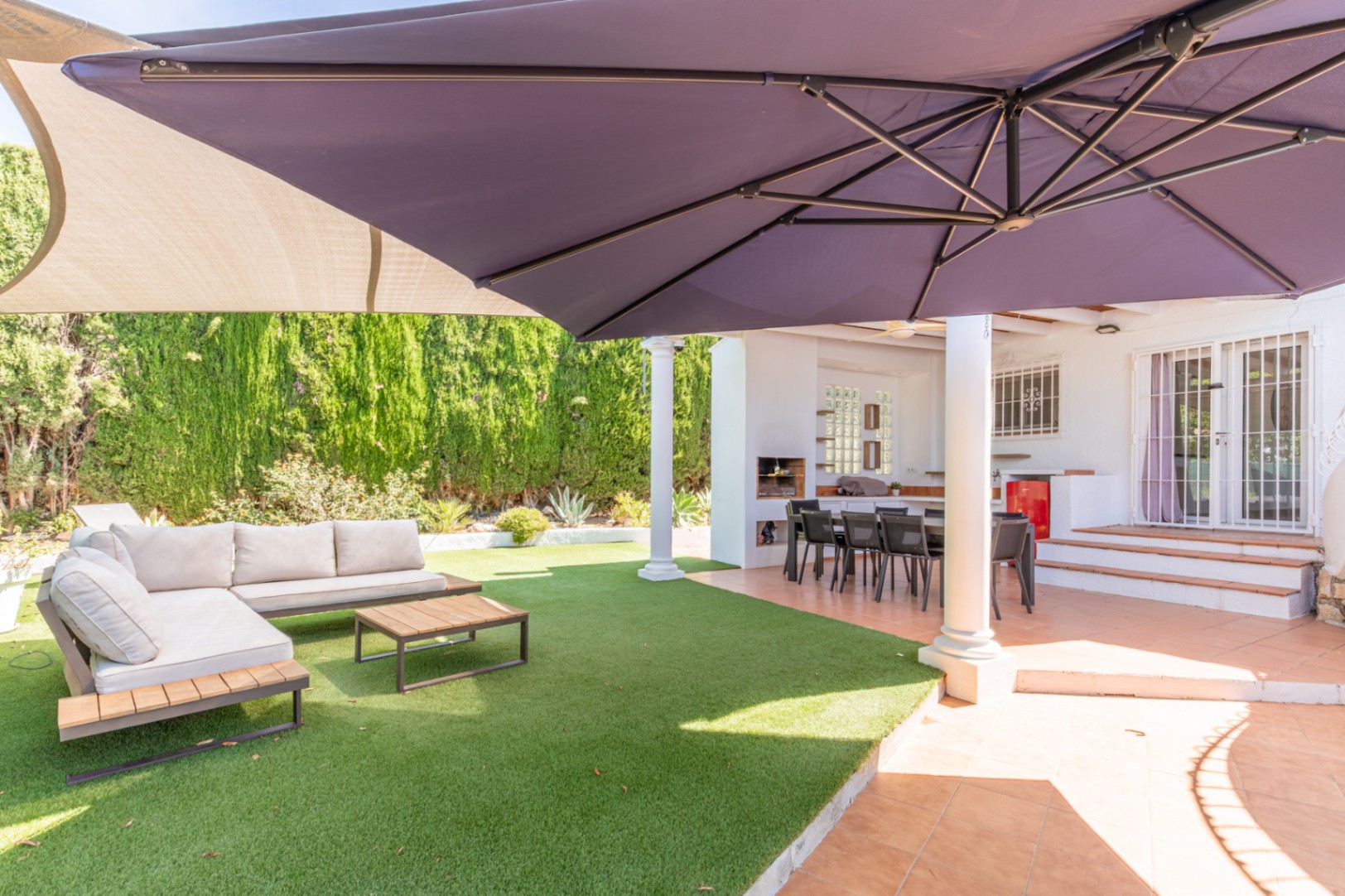 Terraza exterior cubierta con comedor y zona de barbacoa integrada, junto a un césped artificial con mobiliario lounge y una gran sombrilla. Terraza exterior cubierta con comedor y zona de barbacoa integrada, junto a un césped artificial con mobiliario lounge y una gran sombrilla.