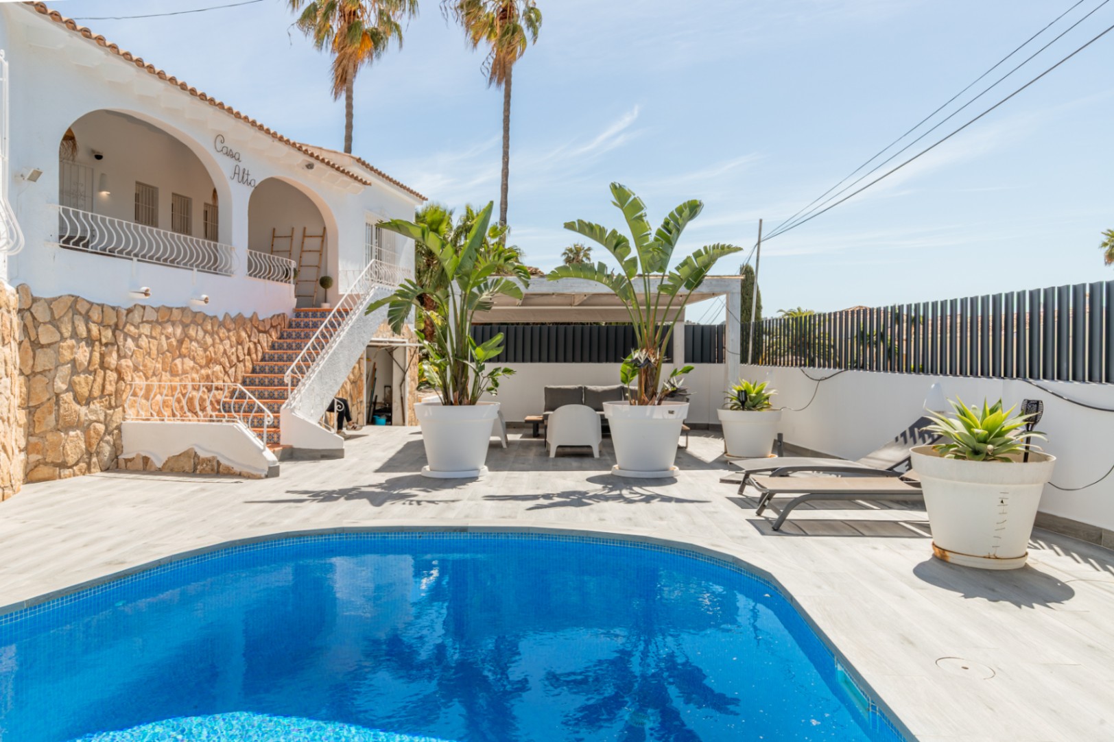 Vista exterior de la villa 'Casa Alta' con piscina de mosaico azul. La terraza de lamas claras rodea la piscina, junto a una escalera exterior con azulejos decorativos y base de piedra. Vista exterior de la villa 'Casa Alta' con piscina de mosaico azul. La terraza de lamas claras rodea la piscina, junto a una escalera exterior con azulejos decorativos y base de piedra.