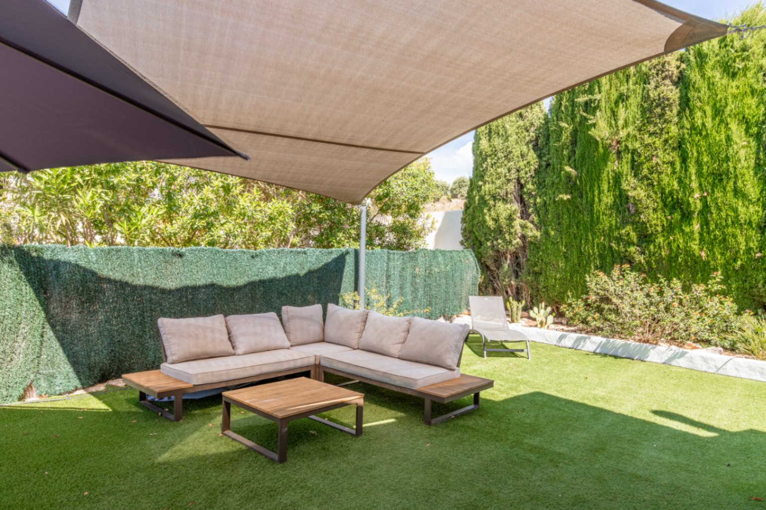 Zona de estar exterior sombreada con sofá en L de cojines claros y mesa de centro de madera sobre césped artificial. Setos altos y árboles ofrecen privacidad bajo una vela de sombra beige. Zona de estar exterior sombreada con sofá en L de cojines claros y mesa de centro de madera sobre césped artificial. Setos altos y árboles ofrecen privacidad bajo una vela de sombra beige.