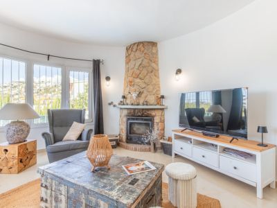Acogedor salón con chimenea revestida de piedra y amplios ventanales curvos que ofrecen vistas a las colinas. Incluye una mesa de centro rústica y un mueble de TV moderno.