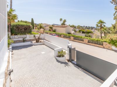 Amplia zona de aparcamiento pavimentada con adoquines en espiga y una moderna puerta corredera gris oscuro. Vistas a la zona residencial, vegetación mediterránea y el mar a lo lejos.