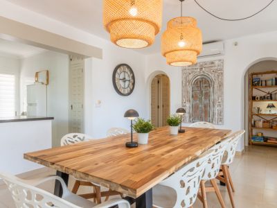 Amplio comedor con mesa de madera maciza, sillas blancas modernas y dos lámparas colgantes de mimbre. Arcos conectan con la cocina y un rincón de lectura con estantería.
