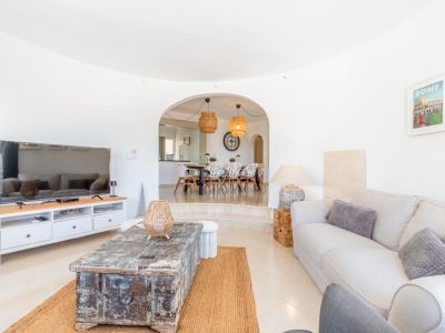 Amplio salón con suelo de baldosas claras, sofá confortable y mesa de centro rústica sobre alfombra de yute. Un arco conduce al comedor elevado con asientos modernos y lámparas colgantes de ratán.