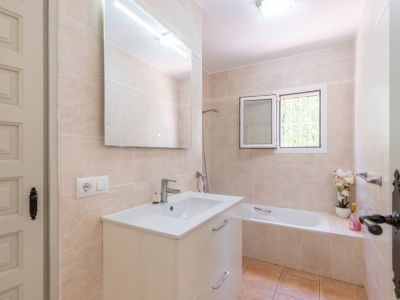 Baño con azulejos beige claro, un mueble de lavabo blanco con espejo grande e iluminación superior. Dispone de bañera y ventana con luz natural.
