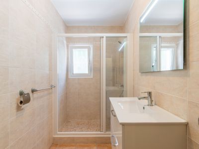 Baño moderno con azulejos beige, lavabo blanco, espejo con iluminación LED y ducha con mampara de cristal, suelo de mosaico y ventana.
