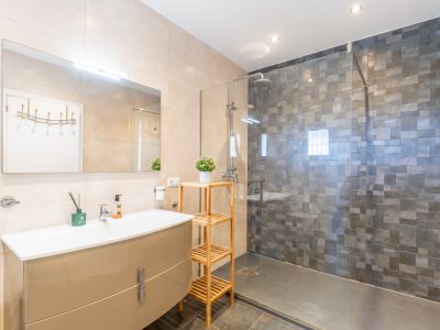 Baño moderno con lavabo de mueble beige brillante, espejo iluminado y ducha de obra con mampara de cristal y azulejos mosaico grises. Suelos oscuros y paredes claras.
