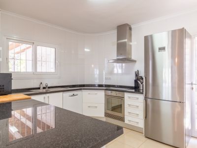 Cocina equipada con armarios blancos, encimeras de granito oscuro y electrodomésticos de acero inoxidable, incluyendo nevera y horno integrados. Ventana con rejas de seguridad que aporta luz natural.
