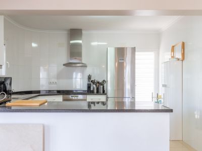 Cocina moderna y luminosa con paredes de azulejos blancos, encimeras de granito oscuro y electrodomésticos de acero inoxidable, incluyendo nevera y campana extractora. Dispone de una barra funcional.