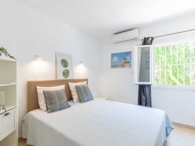 Dormitorio con cama doble, cabecero textil beige, aire acondicionado y ventana con reja de seguridad, ofreciendo vistas a la vegetación exterior.