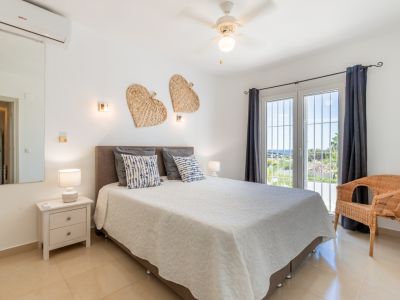 Dormitorio con cama doble, suelo de baldosas claras y puerta al balcón con cortinas oscuras. Disfrute de luz natural, ventilador de techo, aire acondicionado y vistas lejanas al mar.