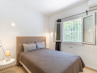 Dormitorio luminoso con cama de cabecero tapizado, gran ventana con rejas de seguridad y vistas a la vegetación, además de aire acondicionado.