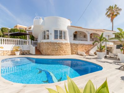 Exterior de villa blanca con piscina privada de mosaico azul y delfín. Destaca la base de piedra y una sección arquitectónica redonda con ventanas.