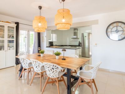 Luminoso comedor con una gran mesa de madera, ocho sillas blancas de diseño y dos lámparas colgantes de mimbre. Se conecta fluidamente a una cocina moderna y equipada con electrodomésticos de acero.